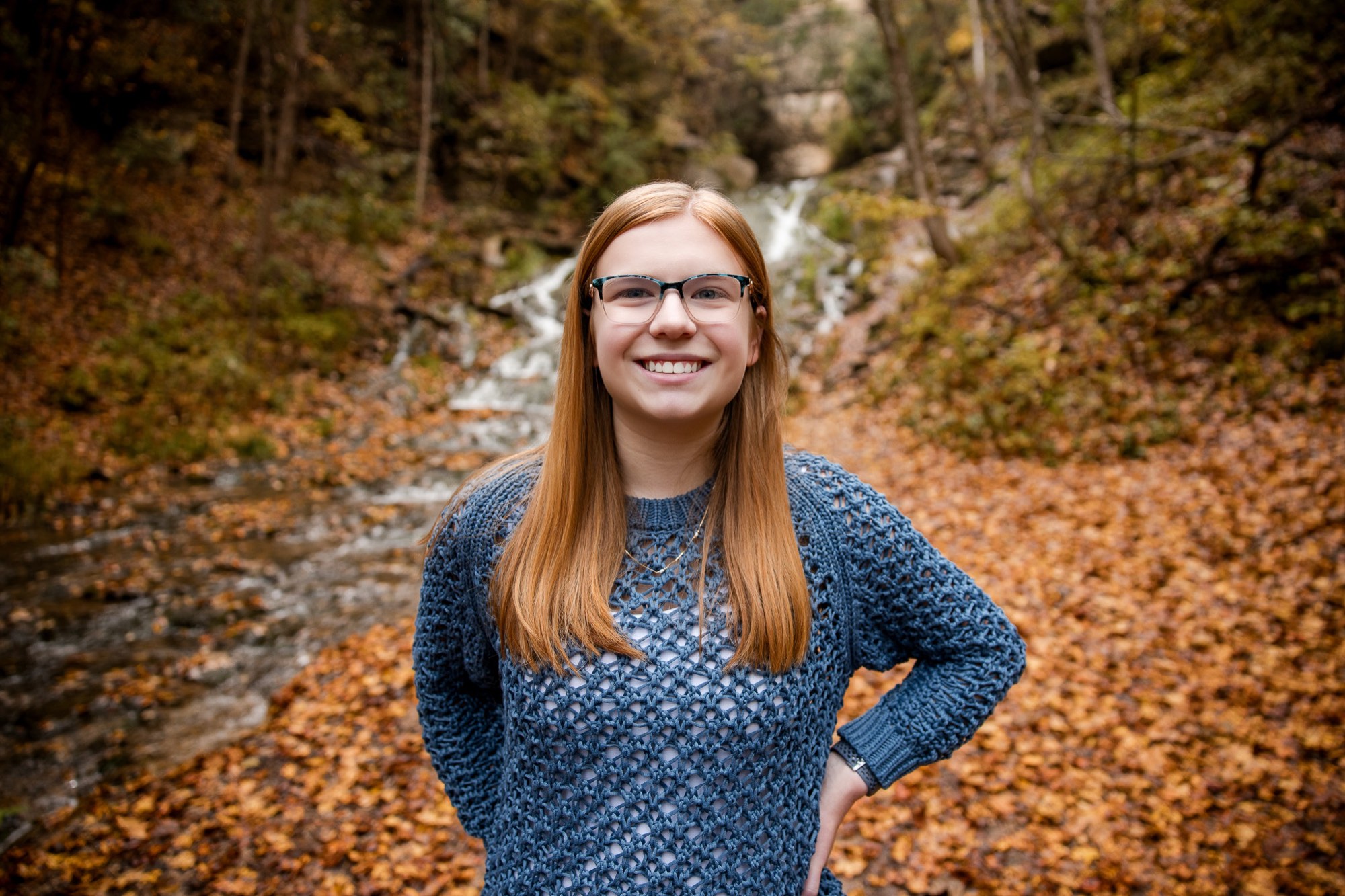 Hannah smiling in a blue sweater in front of a waterfall and fallen leaves