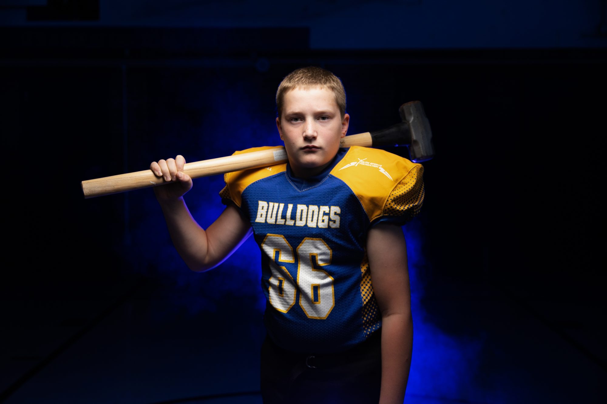 MFL MarMac Youth Sports Foundation football player, #66 Bulldogs, posed with a sledgehammer over his shoulder under blue stage lighting