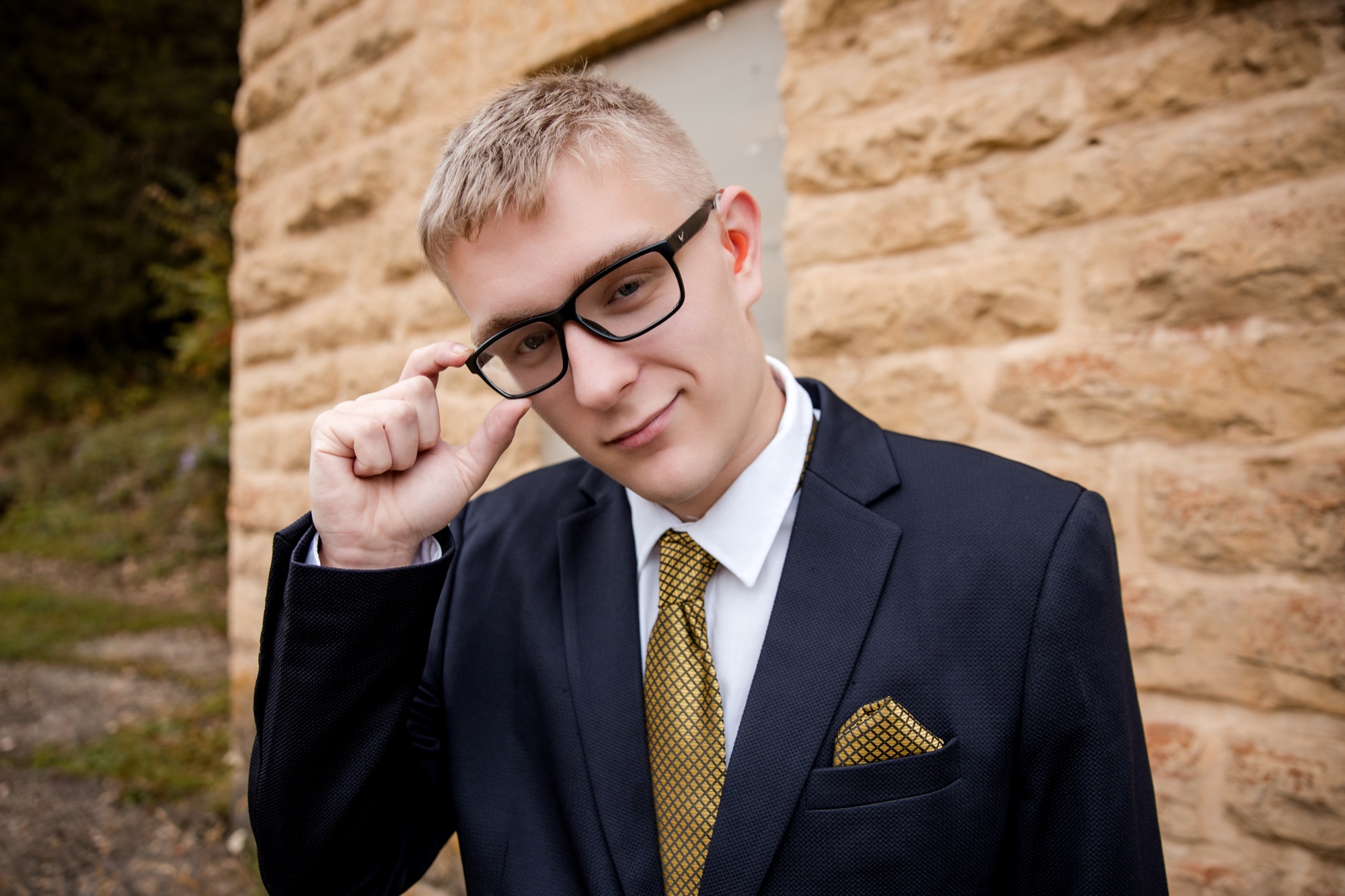 Tucker adjusting his glasses in a suit against a limestone wall